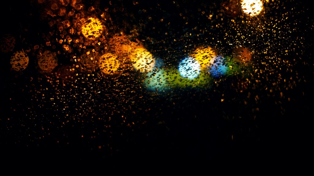 Raindrops on a window with blurred, colorful streetlights in the background create a bokeh effect. The lights, predominantly yellow, orange, blue, and green, contrast against the dark night. This scene resembles a sample processing moment where NZO Cloud adapts to demand spikes seamlessly.