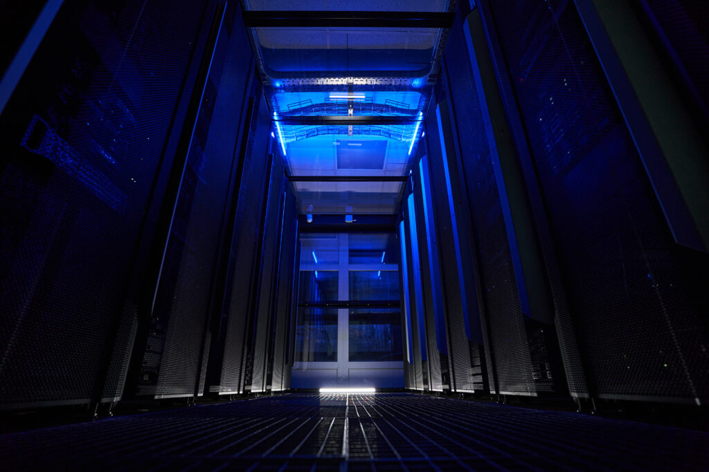 A dimly lit server room with a blue glow, showcasing tall server racks on both sides and a mesh floor. Overhead, cabling and ventilation are visible, illuminated by the blue lighting that creates a futuristic ambiance reminiscent of cloud computing infrastructure.