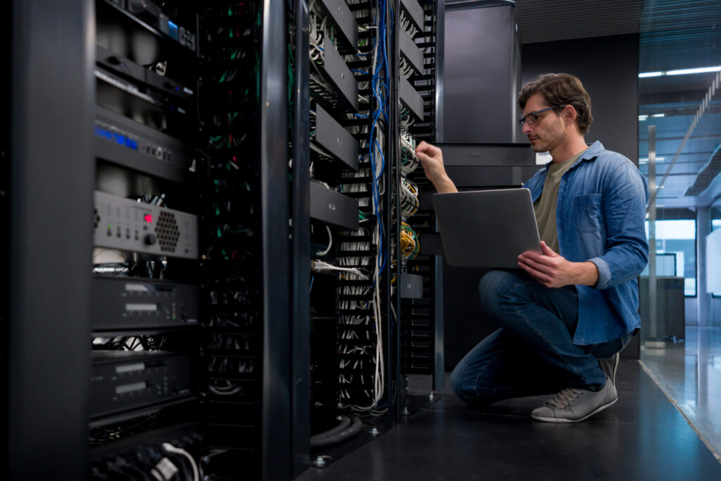 A man wearing glasses and a denim shirt is kneeling in front of a server rack, holding a laptop and inspecting cables in a data center. Surrounded by rows of server equipment and networking hardware, he focuses on solutions for cloud cost optimization amid the challenges.