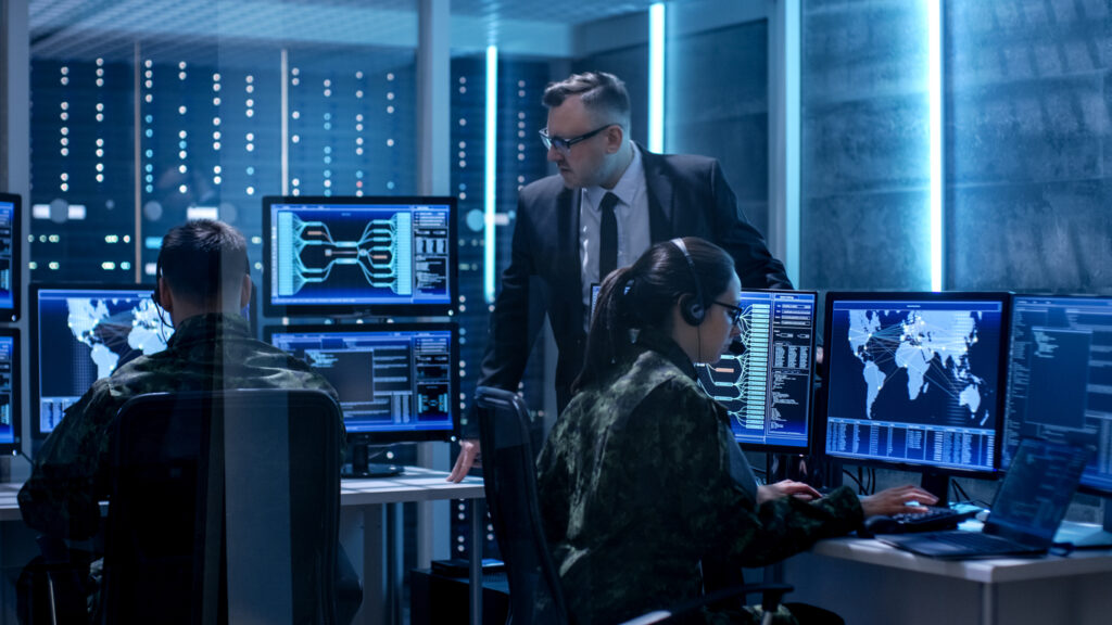 In a control room with multiple screens displaying maps and data, two people in military uniforms work on computers while a person in a suit oversees their activities. The scene is illuminated by the glow of monitors and dim, ambient lighting.